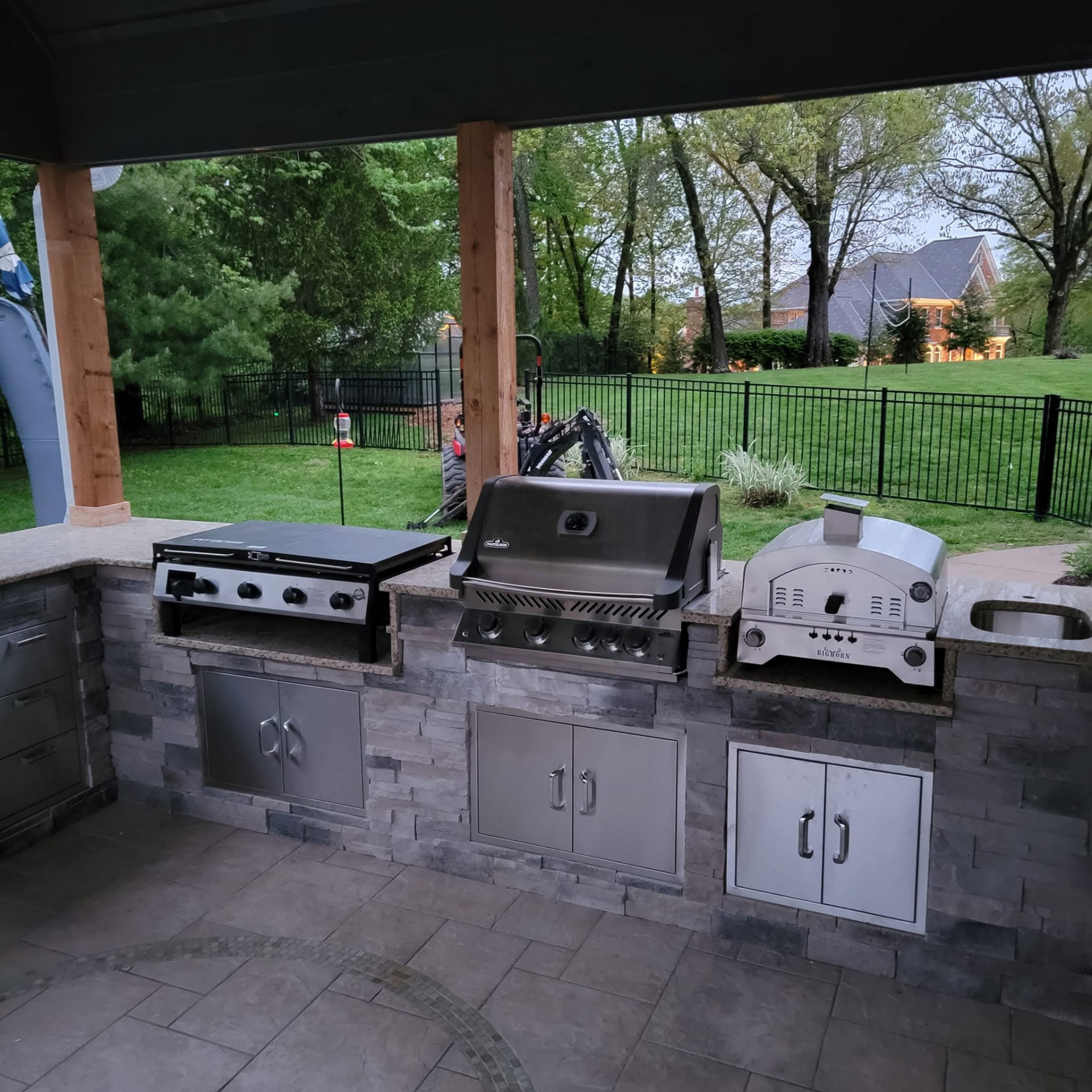 Full outdoor cooking station — gas grill, Blackstone griddle, pizza oven, and a sink, all stainless under the covered roof by B&R Construction